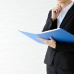 ファイルを片手に顎へ手を当てて考えているスーツ姿の女性