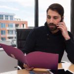 オフィスでファイルを片手に電話で話している男性