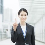 屋外にて笑顔で人差し指を立てているスーツ姿の女性