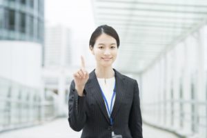 屋外にて笑顔で人差し指を立てているスーツ姿の女性